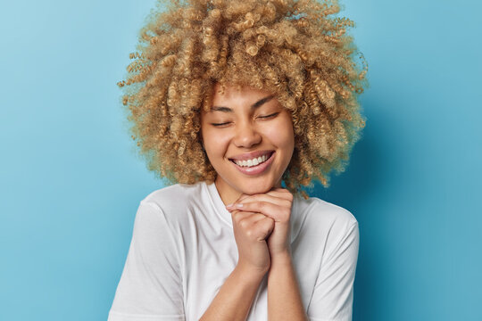 Cheerful curly haired woman keeps hands under chin has closed eyes smiles broadly expresses sincere positive emotions and feelings dressed casually isolated over blue background. Happiness concept