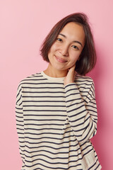 Fototapeta premium Vertical shot of pleased millennial Asian woman with dark hair keeps hand on neck smiles gently dressed in casual striped jumper tilts head isolated over pink background. Positive human feelings
