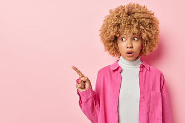 Shocked excited woman with curly hair points finger on left shows blank space for your promo opens mouth from amazement wears turtleneck and shirt isolated over pink background. Omg look there