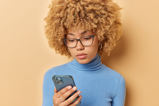 Serious looking confident woman with curly blonde hair holds smartphone makes appointment online focused at cellular display wears transparent eyeglasses and blue turtleneck isolated over beige wall