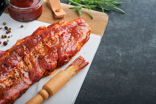 Pork ribs. Raw meat. Raw pork ribs in marinade with spices, rosemary, tomato sauce and garlic on white marble stand on black stone table background. Barbecue concept. Selective focus. Mock up.