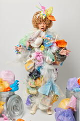 Photo of shocked curly woman collects garbage works as volunteer cleans up waste for recycle makes costume from rubbish isolated over grey background. Global problem and contamination concept