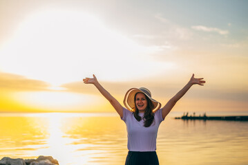 portrait of a happy woman on a journey in a hat in the summer at sunset on the seashore with her hands up