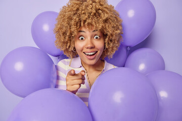 Fototapeta na wymiar Surprised amazed curly haired woman points index finger directly at camera notices something strange prepares for holiday or party surrounded by purple inflated balloons. Happy birthday concept