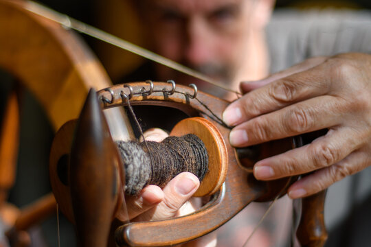 Skilled tradesman works to repair and to intricately fettle the mechanism of an old wooden spinning wheel. Selective focus to accentuate the task of repair and service. Contrasting lighting