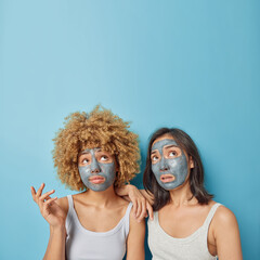 Vertical shot of confused two mixed race women look overhead apply clay facial masks for skin treatment dressed in casual t shirts isolated over blue background empty space for your promotion