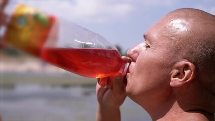 Bald Wrinkly Man Greedily Drinks Juice, Drink, Colored Water Against the Sky. Man drinks a red drink from a plastic bottle in the rays, glare of sunlight. Thirst in nature, on the beach. Summertime.