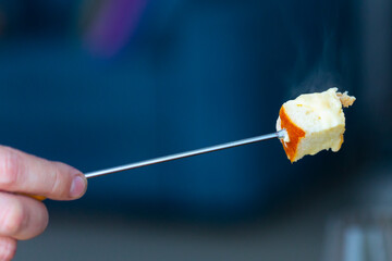 Piece of traditional French cheese fondue with hot steam on fondue fork in hand of man  