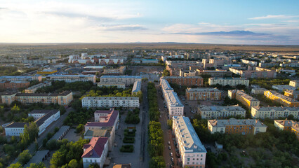 A small town of Balkhash with green trees. Five-storey houses of different colors. There is a green alley and a road with cars in the center of city. Dark clouds and mountains are visible in distance