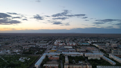 Dark clouds on the horizon. Sunset over a small town. Top view from a drone. Low colored houses are standing. Cars are driving on the road. Lights and house lights are on. The steppe is far away