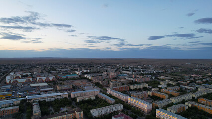 Dark clouds on the horizon. Sunset over a small town. Top view from a drone. Low colored houses are standing. Cars are driving on the road. Lights and house lights are on. The steppe is far away