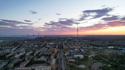 Orange sunset over a small town. Top view from a drone. Blue-purple clouds against background of houses, a smoking factory and a TV tower. Cars drive. There is a bright light at the metal drain plant