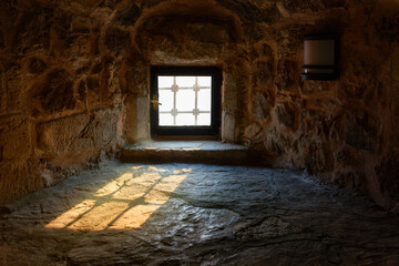 Bright sunlight enters the dark stone room through a barred window.