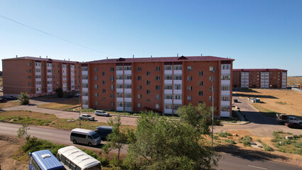 A new area on the outskirts of the small town of Balkhash. Brickwork of the walls. A five-storey house with plastic windows and balconies. There are cars on the road. Green trees are growing. Blue sky