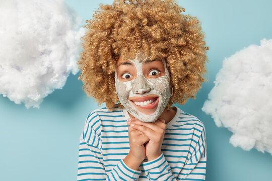 Headshot of cheerful young European woman with curly hair keeps hands under chin bites lips looks gladfully at camera applies clay mask on face for skin treatment isolated over blue background
