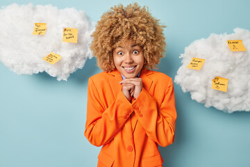 Positive woman dressed in elegant orange jacket keeps hands under chin smiles happily uses reminder notes writes list to do poses against blue backgrouns white fluffy clouds overhead. Multitasking