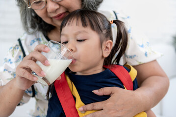 Asian preschool girl, age 3-year-old, drinking milk from a glass which Grandma is holding for her to drink, with white background, to milk for children health care concept.