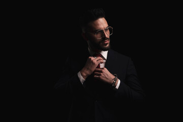 cool elegant fashion man in black suit looking to side and fixing tie