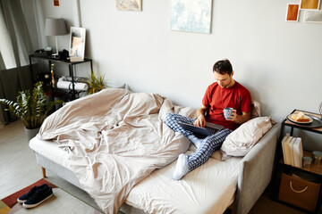 Obraz premium High angle view of young man sitting on bed and browsing in laptop with cup of coffee in the morning