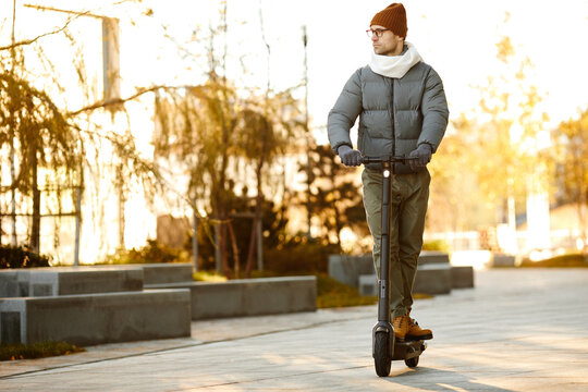 Young man in warm clothing riding on scooter along the street in autumn day