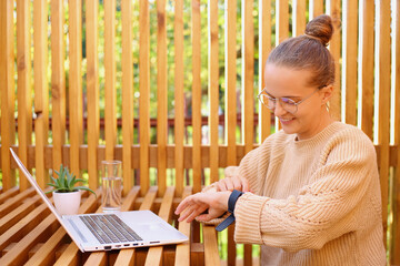 Portrait of smiling satisfied dark haired female wearing warm jumper sitting at table and working on laptop, checking her smartwatch with happy facial expression.