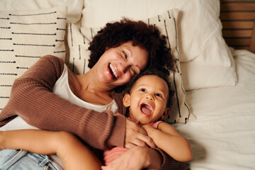 Happy biracial mother tickling daughter and laughing on bed at home - obrazy, fototapety, plakaty