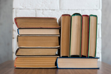 A stack of hardcover books on a wooden table against a white brick wall