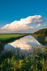 Dissipating storm cloud is reflected in the calm water of a canal on a serene summer evening