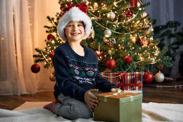Obraz na płótnie Canvas A happy boy in a santa claus hat opens a box with a Christmas present next to a Christmas tree in garlands. Happy New Year and Christmas holiday