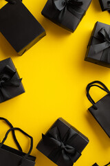 Black friday sale concept. Top view vertical photo of black gift boxes with ribbon bows and paper bags on isolated yellow background with empty space in the middle