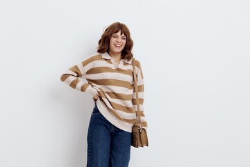 a joyful woman stands on a white background in a striped sweater and with a bag on her shoulder put her hand on the belt pulling the sweater up a little, putting her other hand on the bag