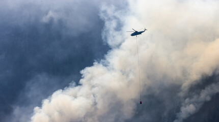Fototapeta na wymiar Wildfire Service Helicopter flying over BC Forest Fire and Smoke on the mountain near Hope during a hot sunny summer day. British Columbia, Canada. Natural Disaster