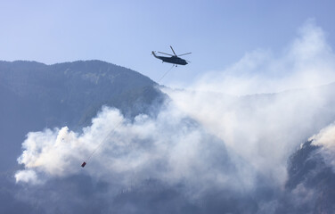 Fototapeta premium Wildfire Service Helicopter flying over BC Forest Fire and Smoke on the mountain near Hope during a hot sunny summer day. British Columbia, Canada. Natural Disaster