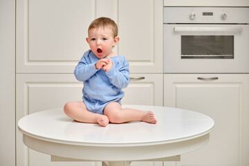 Toddler baby climbed on the kitchen white table. A child in danger climbed to the top, risking falling off the furniture. Kid aged one year and two months