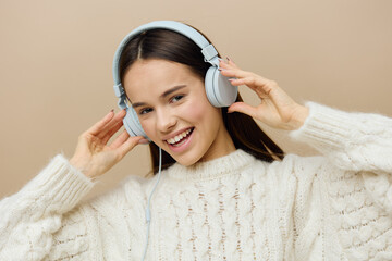 close horizontal photo of a woman in a white sweater listening to music in her wired headphones smiling broadly with happiness and holding them with her hands while looking at the camera with delight