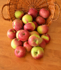 Fresh red and green apples falling from a wicker basket on a wooden table.