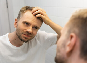 Obraz premium Middle aged caucasian white man with a short beard looks at his hair in the mirror in the bathroom and worried about balding. The concept of the problem of male hair loss, early baldness and alopecia