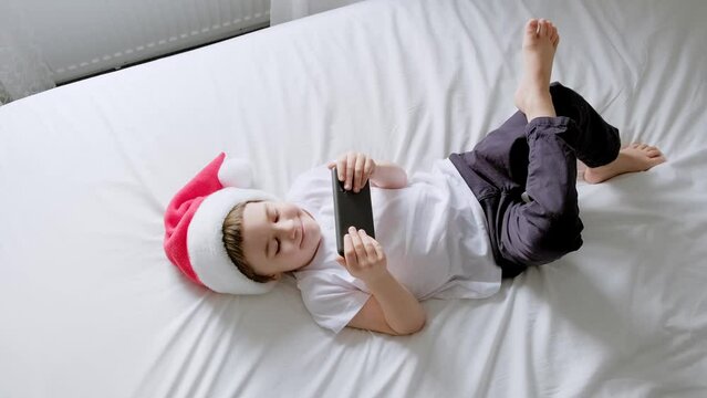 5 year boy play smartphone video game. Child in Santa Claus hat lie on bed. White home bedroom. Kid use mobile phone for gaming, learning. Spend time speaking with friends. Online education. Close-up.