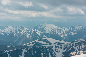 Fototapeta premium Awesome gloomy aerial view to high snow mountain peak in overcast. Atmospheric mountain landscape at high altitude with cloudiness. Alpine scenery with snowy mountain top in center under cloudy sky.