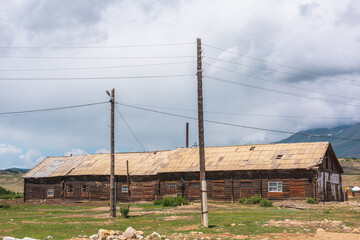Countryside landscape with large long wooden barn with sloping roof among poles with wires in sunlight under cloudy sky at changeable weather in mountains. Sunlit old wide wood house under low clouds.