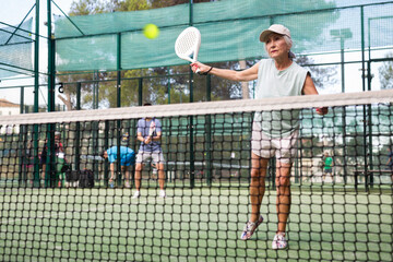 Portrait of active emotional elderly woman playing padel tennis on open court in summer, swinging...