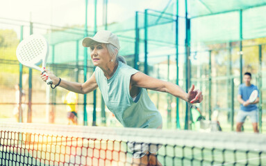 Sporty mature woman padel player hitting ball with a racket on a hard court