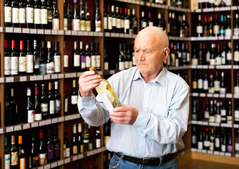 Confident grey-haired elderly man choosing white wine in modern wineshop. High quality photo