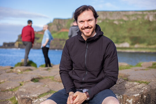 A Caucasian Man In Casual Black Clothes Sitting On Giant's Causeway Columns. Defocused People On Background. County Antrim, Northern Ireland