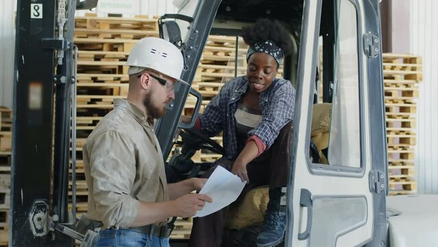 Medium shot of warehouse worker talking to female colleague sitting inside truck lift, discussing transportation and storage of goods
