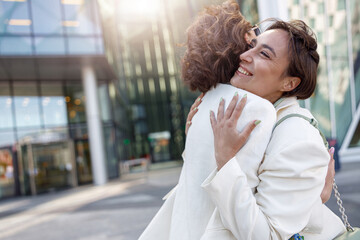 Two emotional happy woman friends in trendy outfits hugging each other on city street