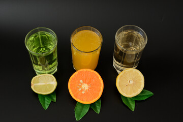 Three glasses with different juices, orange, lemon and lime on a black background, next to the leaves and pieces of ripe citrus fruits.