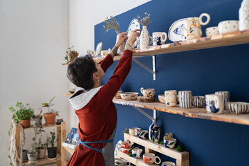Small business female owner putting plate at right place in pottery studio. Self employed girl pottery studio owner tidies up the shelves with handcraft ceramics at her cozy workplace