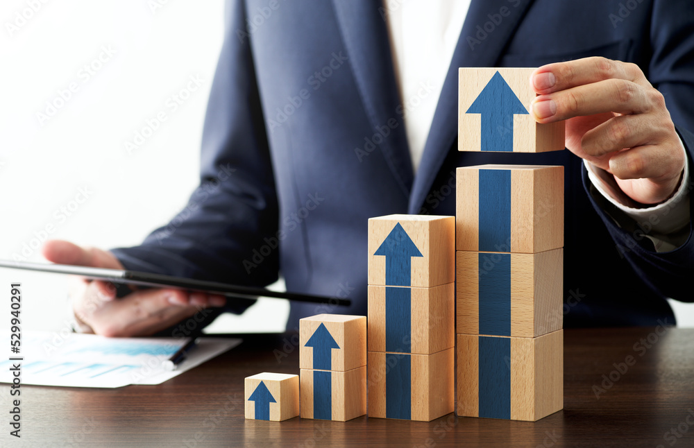 Poster Consultant presenting convincing growth strategy. Analyzing management data and developing project. Man stacking Wood cubes  on desk. Four blue arrows printed on cubes.