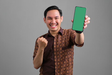 Excited young Asian man wearing batik shirt showing blank screen smartphone and making victory gesture isolated on grey background